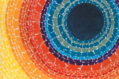 An abstract painting by Alma Thomas featuring concentric, circular rings of vibrant color radiating outward from a large, solid dark blue circle positioned in the upper right. Moving outward from the center, the concentric bands transition through shades of teal, deep blue, purple, bright red, orange, and finally a broad field of sunny yellow that fills the left and bottom edges of the canvas.