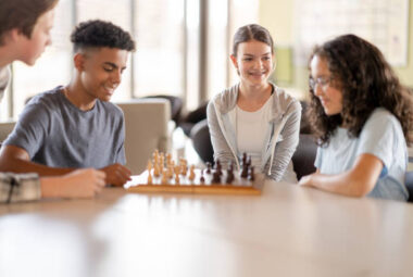 Two teenagers sit at a table playing a game of Chess. They are dressed casually and focused on their next move as they study the board. Two friends are seating next to them watching the game unfold.
