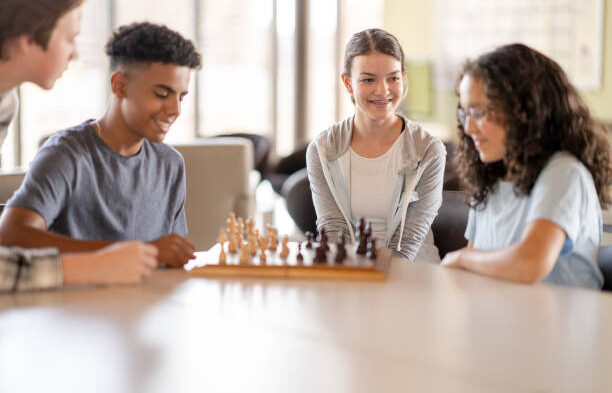 Two teenagers sit at a table playing a game of Chess. They are dressed casually and focused on their next move as they study the board. Two friends are seating next to them watching the game unfold.