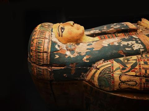 An image of an ancient Egyptian coffin, showcasing a detailed profile view of the face and upper torso carved on the lid. The paint has aged and includes intricate hieroglyphics and symbols.